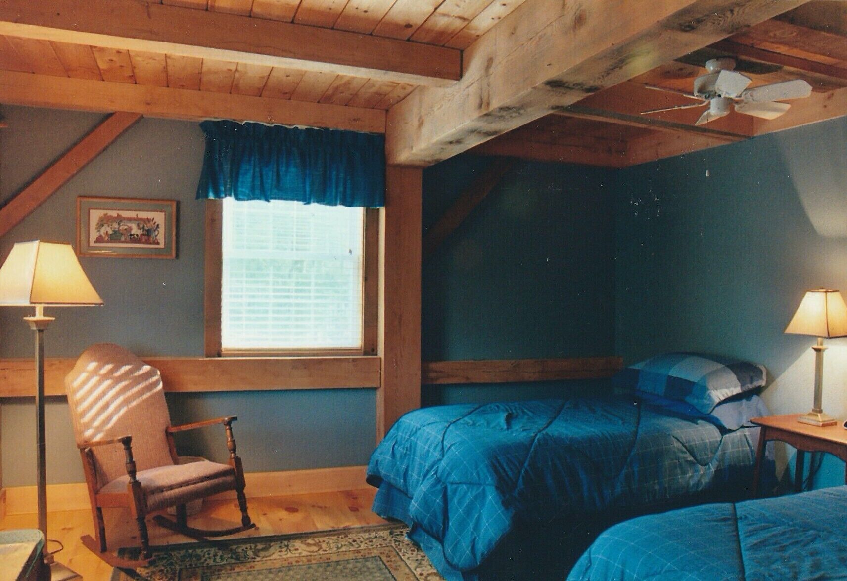 A bedroom with a wooden ceiling, a window with a blue valance, a bed with blue bedding, a rocking chair, and two bedside lamps.