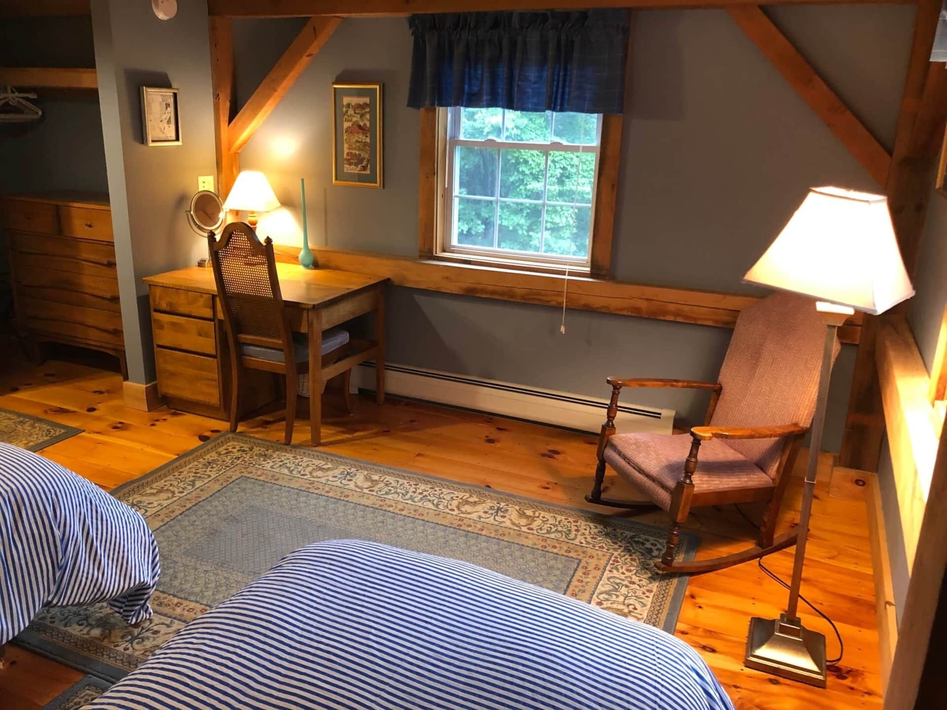 A cozy room with a wooden desk, a rocking chair, a bed with striped bedding, and a window with a view of trees.