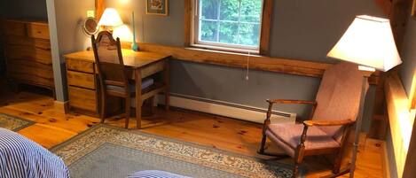 A cozy room with a wooden desk, a rocking chair, a bed with striped bedding, and a window with a view of trees.