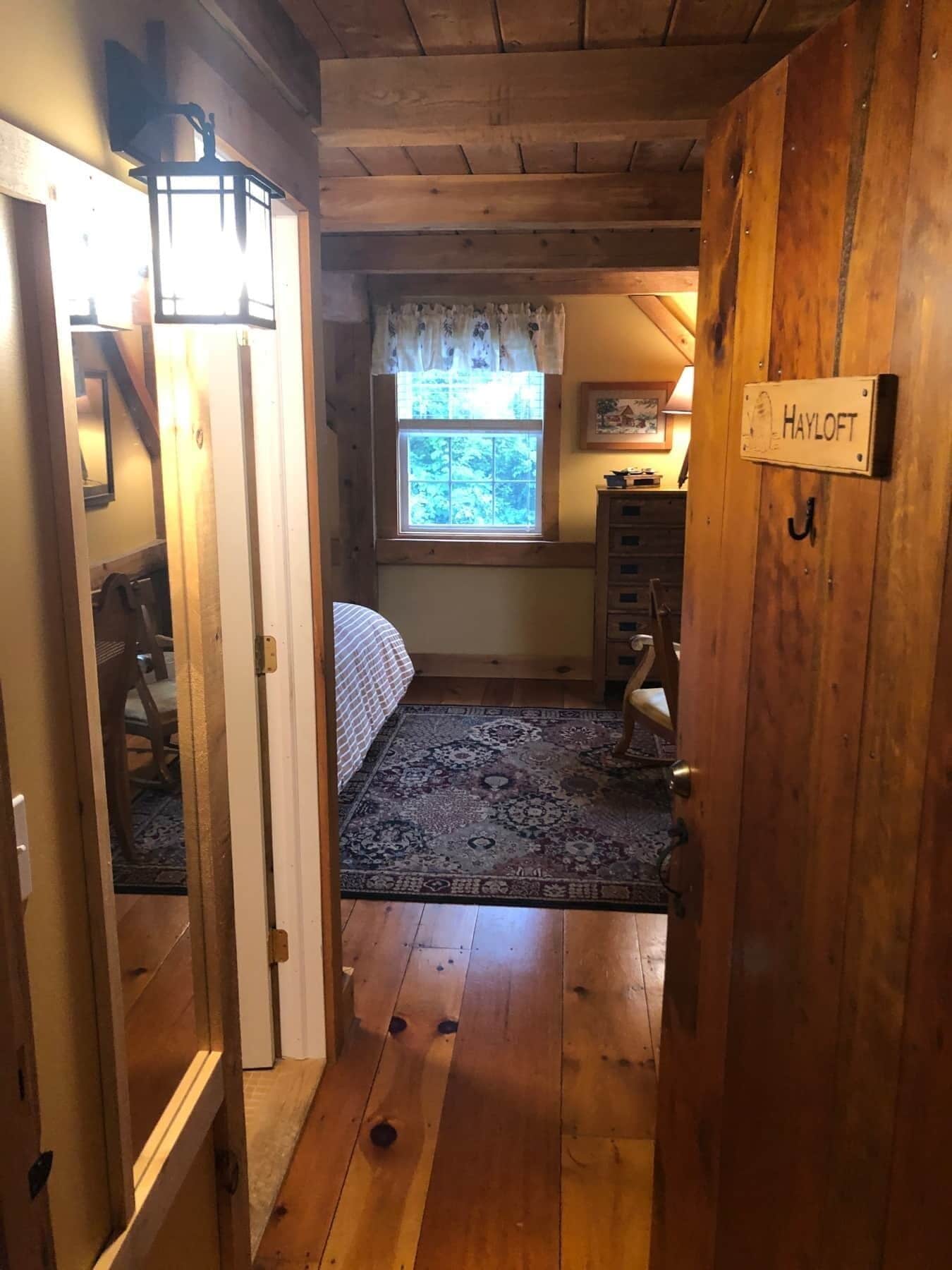 A cozy room with a wooden floor, a bed with a patterned rug, a window with a view of trees, and a sign that reads "HAYLOFT".