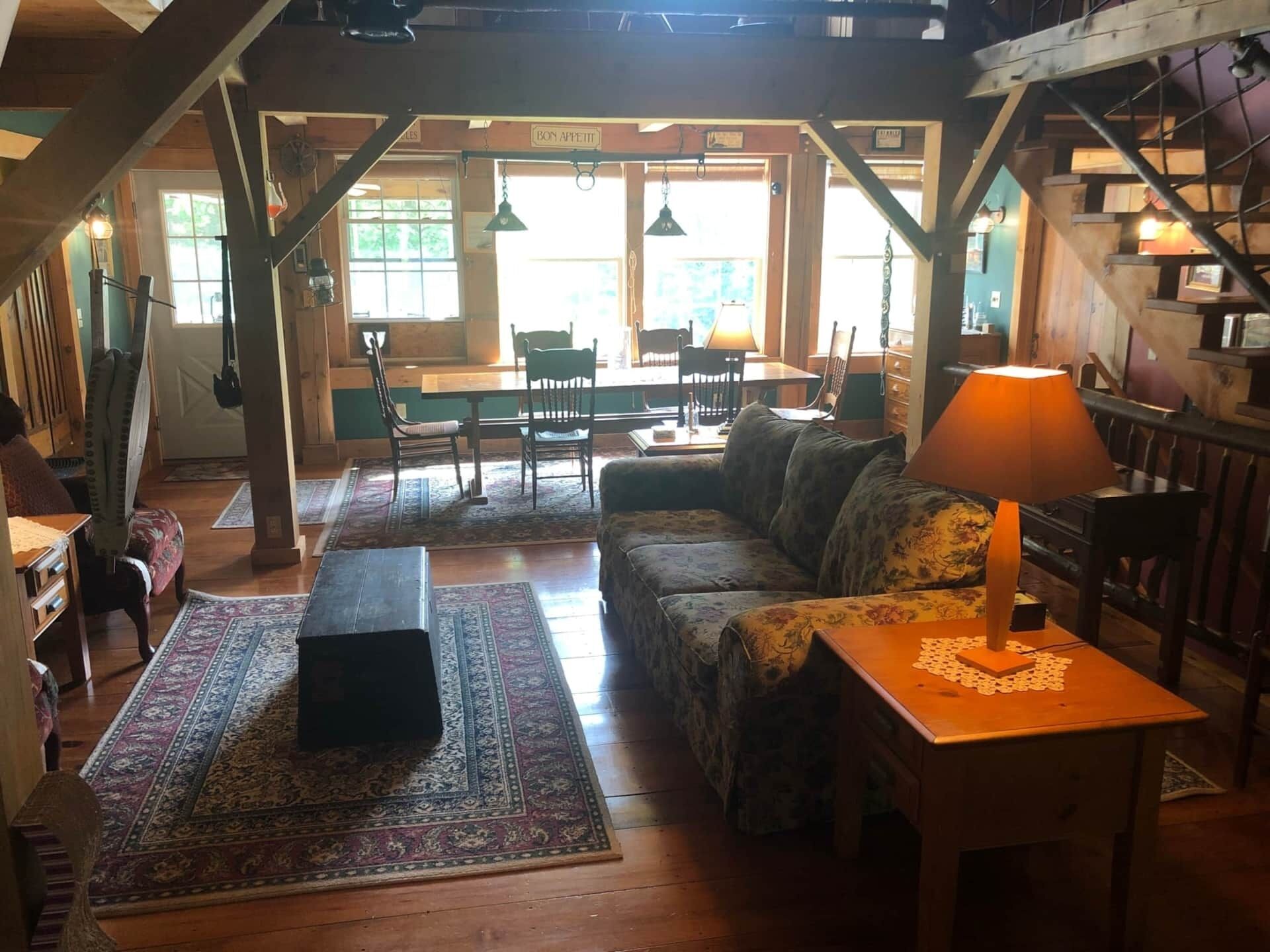 A cozy living room with a wooden staircase, a sofa, a table with a lamp, and a dining area with chairs and a table.