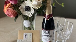 Una botella de jugo de uva roja espumoso Welch's, un ramo de flores, dos copas de vino vacías y una tarjeta con el texto 