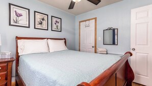 Un dormitorio con una cama grande, cabecera de madera, una mesita de noche, un aparador y un ventilador de techo. Hay tres cuadros con flores enmarcados en la pared, una puerta con espejo y una puerta blanca.