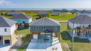 A coastal residential area with houses on stilts, a clear view of the ocean, and a well-maintained lawn. - Beach View Retreat w/ Huge Covered Patio + Games (Bolivar Peninsula)