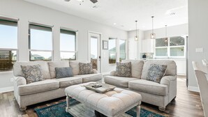 A modern living room with two beige sofas, a matching ottoman, and a ceiling fan. - Beach View Retreat w/ Huge Covered Patio + Games (Bolivar Peninsula)
