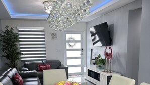 A modern living room with a glass dining table, a chandelier, a sofa, a TV, and a potted plant. - Elegant beautiful house w/pool 10 min from Miami Int. Airport (Hialeah)