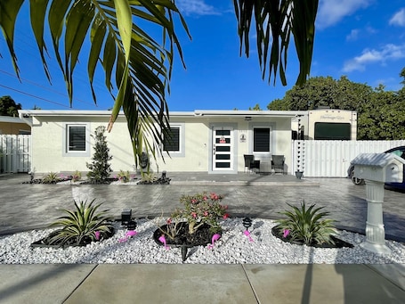 A modern single-story house with a white exterior, a covered patio area, and a small garden with decorative flamingos.