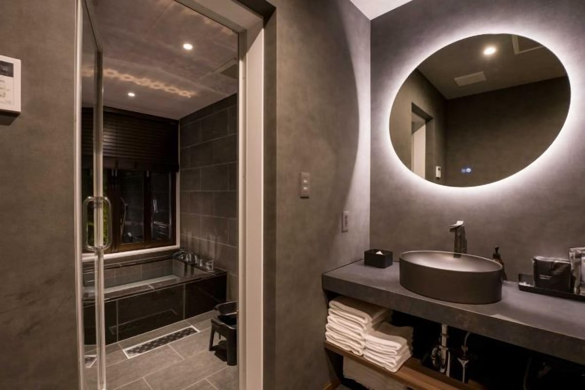 A modern bathroom with a glass-enclosed shower, a round mirror, and a dark-toned sink.