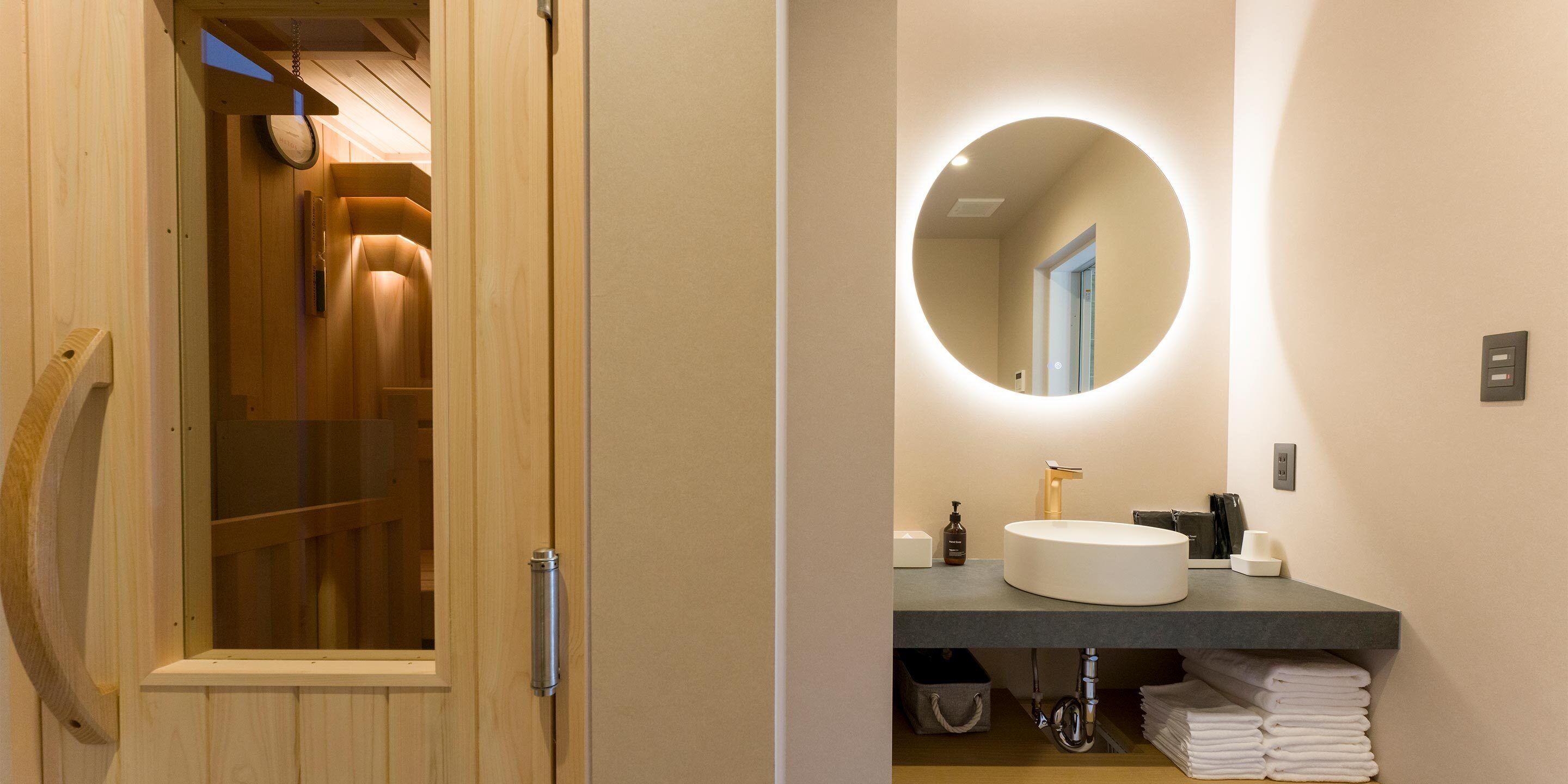 A modern bathroom with a wooden sauna door, a round mirror with a light, a sink, and neatly folded towels.