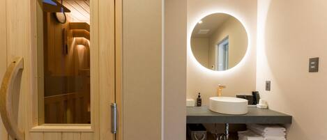 A modern bathroom with a wooden sauna door, a round mirror with a light, a sink, and neatly folded towels.