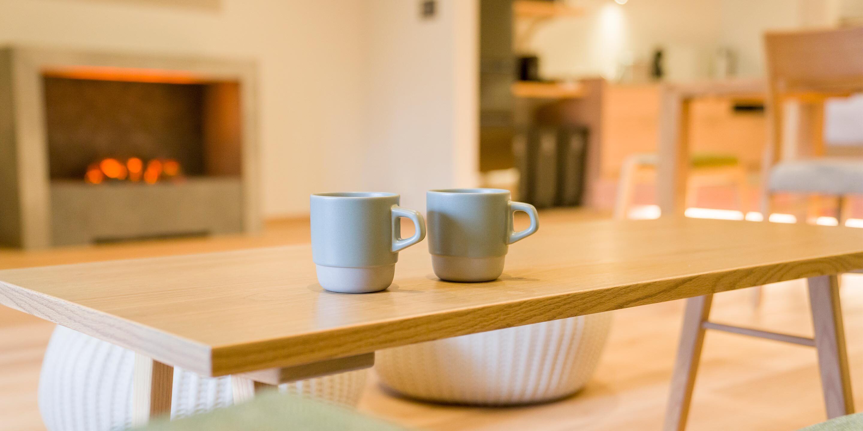 Een houten tafel met twee witte mokken, een witte, beklede bank en een openhaard op de achtergrond.