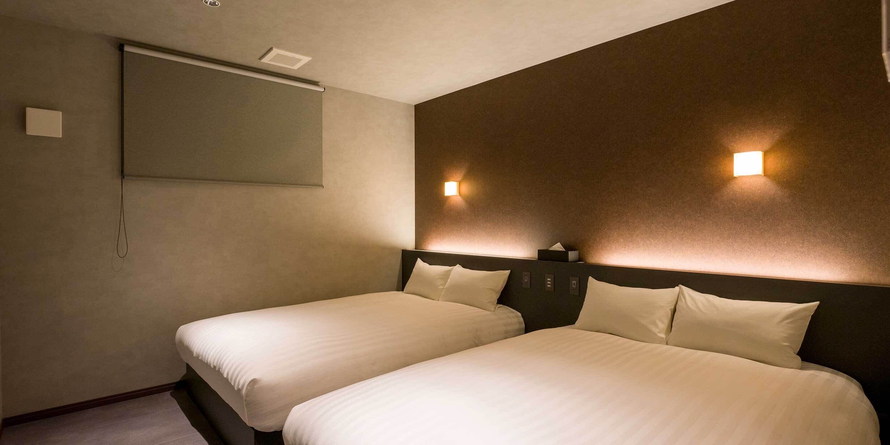 A hotel room with two beds, a ceiling fan, a wall-mounted projector, and two wall sconces.