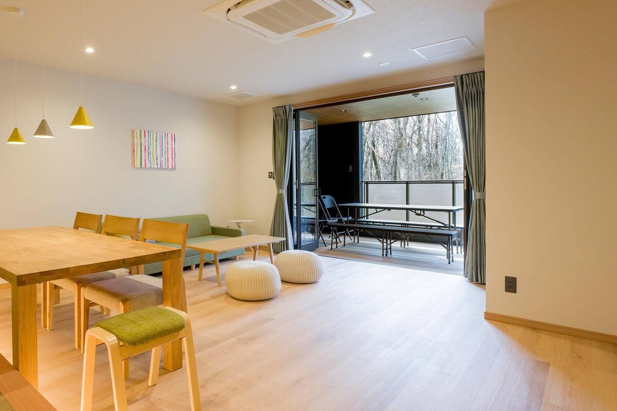 A modern living room with a wooden dining table, green cushioned chairs, a sofa, and a large window with curtains.