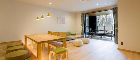 A modern living room with a wooden dining table, green cushioned chairs, a sofa, and a large window with curtains.