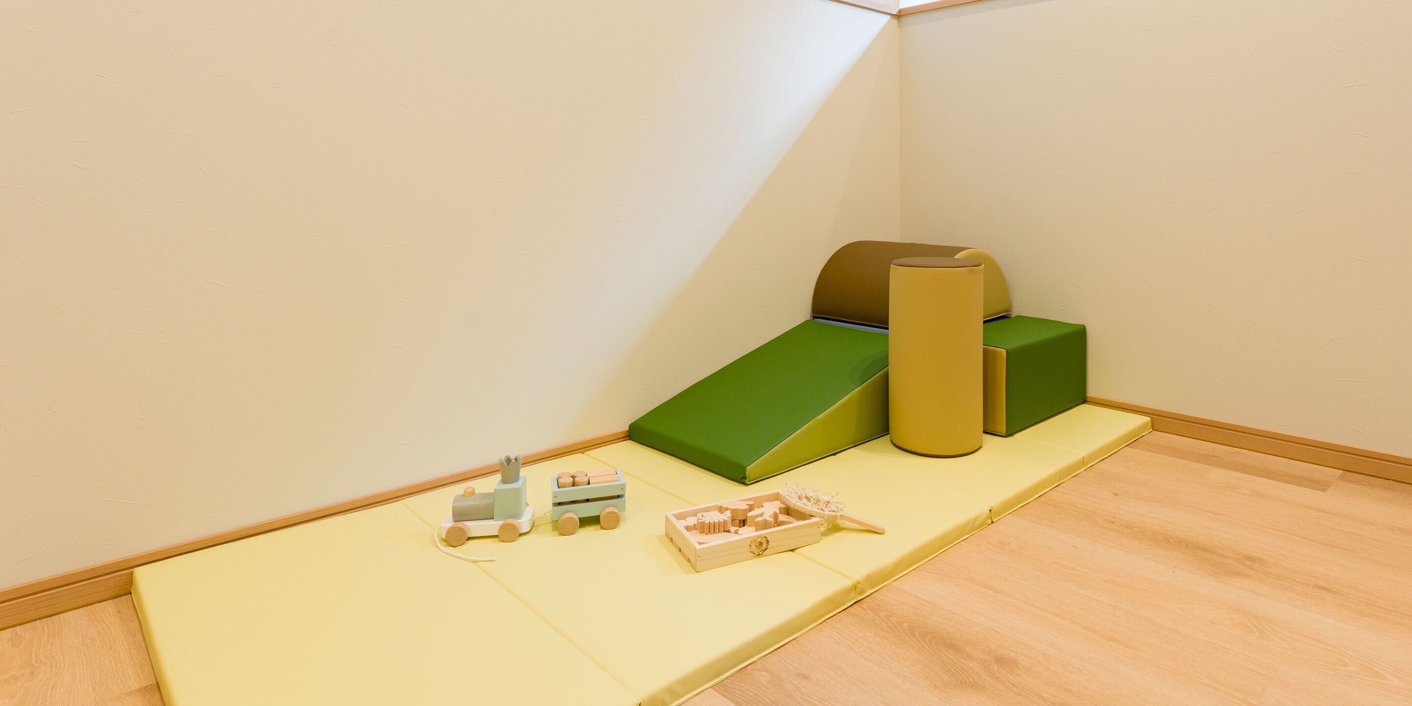 A corner of a room with a wooden floor, a green cushioned bench, a yellow cylindrical object, and a small wooden train set.