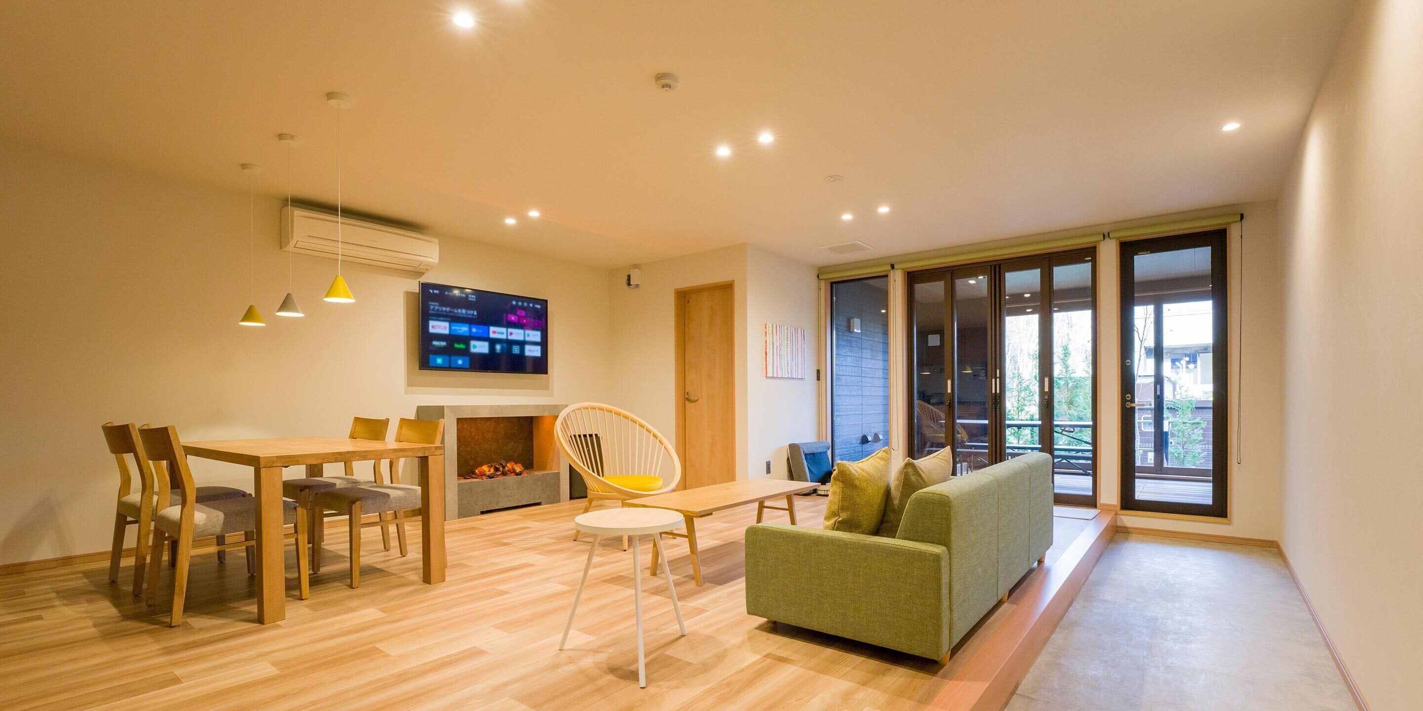 A modern living room with a wooden dining table, chairs, a sofa, and a flat-screen TV mounted on the wall.
