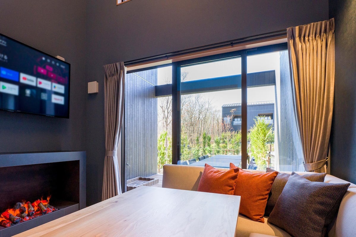 A modern room with a flat-screen TV mounted on the wall, a fireplace, a sofa with cushions, and a large window with curtains.