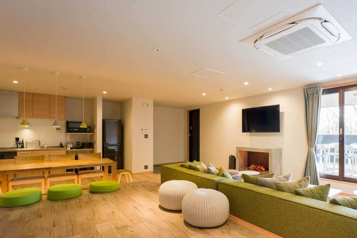 A modern living room with a green sofa, ottomans, a fireplace, and a large window overlooking an outdoor area.