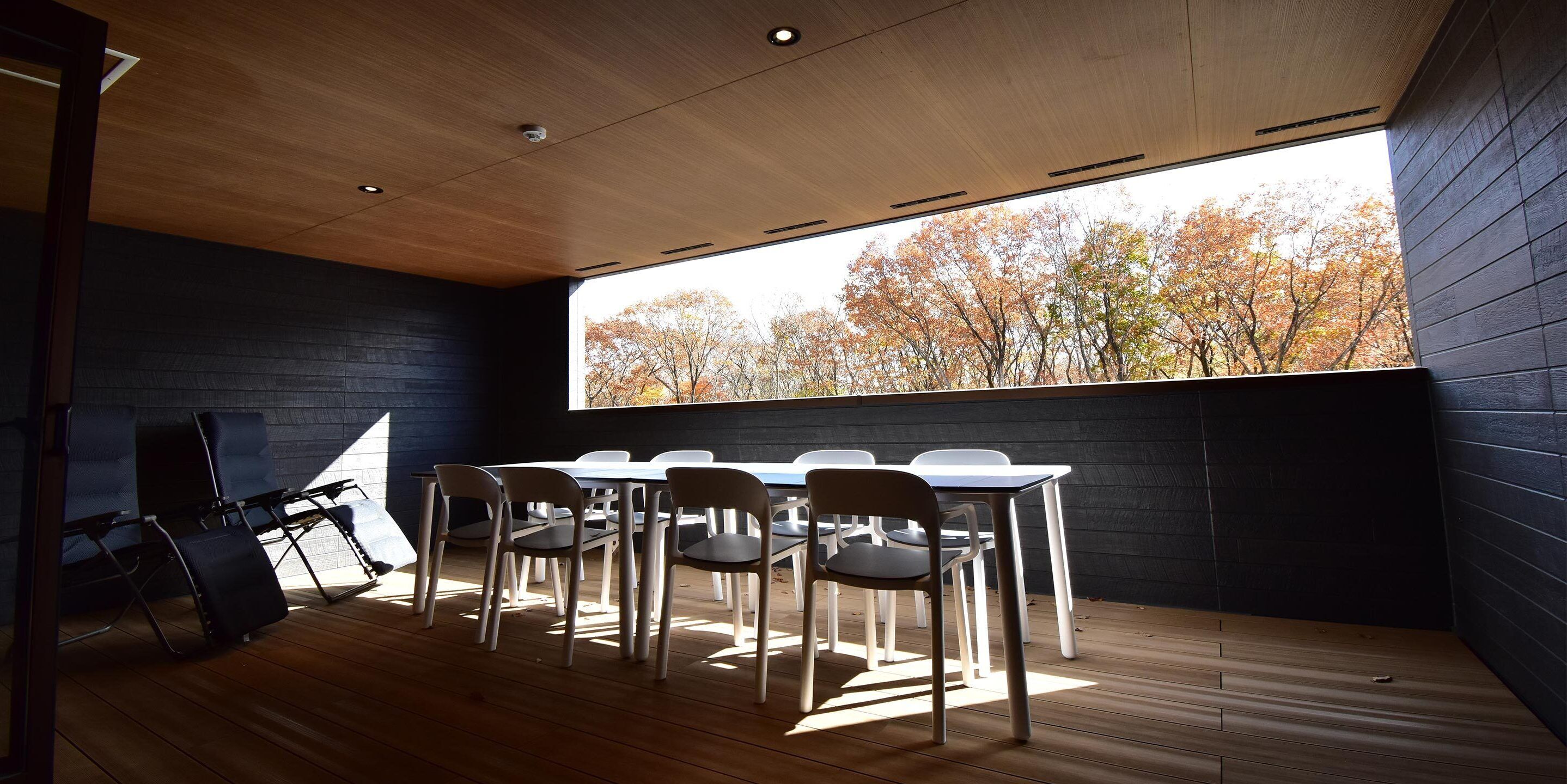 A modern meeting room with a large table and chairs, wooden flooring, and a view of autumn trees through a large window.