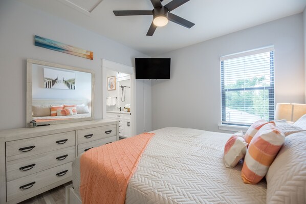 Une chambre à coucher comprenant un lit, une commode, un ventilateur de plafond, un téléviseur et une fenêtre avec des stores.