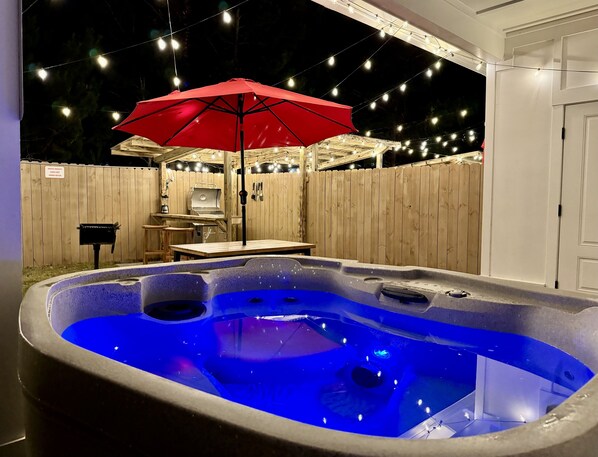 Uma banheira de hidromassagem com iluminação azul, um guarda-sol vermelho e um deck de madeira com luzinhas.