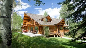 Une cabane en rondins avec une terrasse, entourée d’arbres et d’une pelouse.