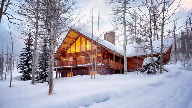 Une cabane en rondins dotée d’une grande façade vitrée et d’un porche couvert, entourée de neige et d’arbres.