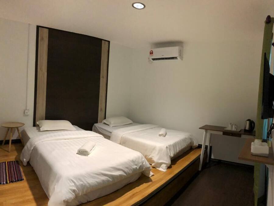 A hotel room with two single beds, a small table, a chair, an air conditioning unit, and a kitchenette.