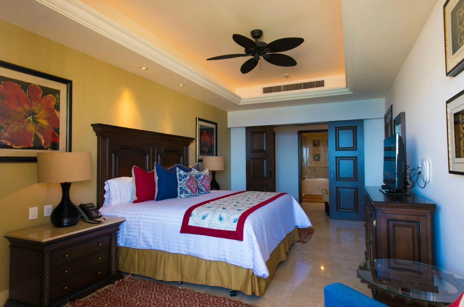 A hotel bedroom with a bed, bedside lamps, a ceiling fan, and a television on a wooden cabinet.