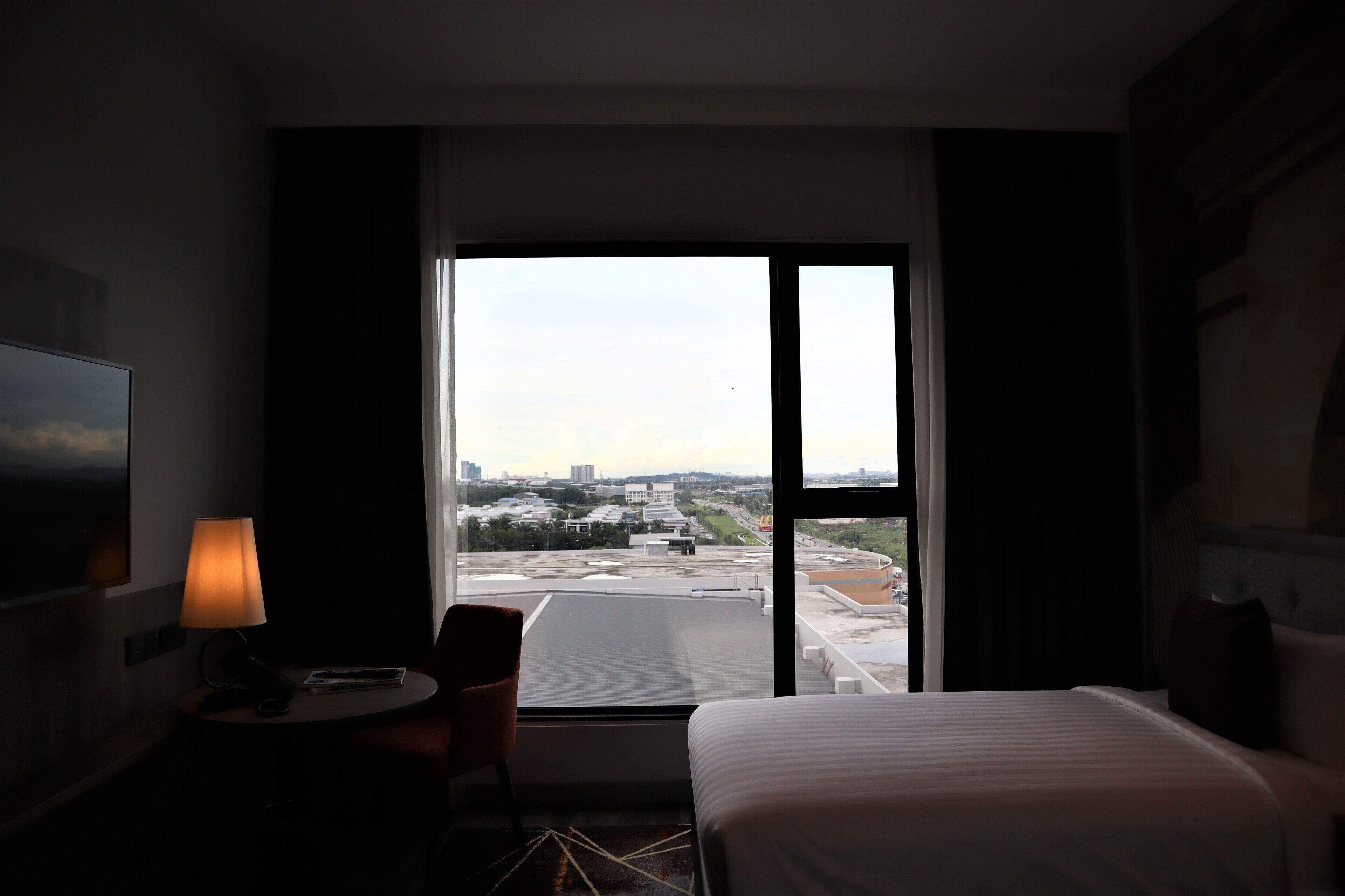 Une chambre d’hôtel avec un lit, un bureau doté d’une lampe, une chaise et une grande fenêtre donnant sur la ville.