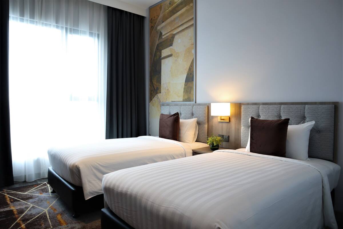 Ein Hotelzimmer mit zwei Betten, einem großen Fenster mit Vorhängen, einem Gemälde an der Wand und einem Nachttisch mit Lampe.