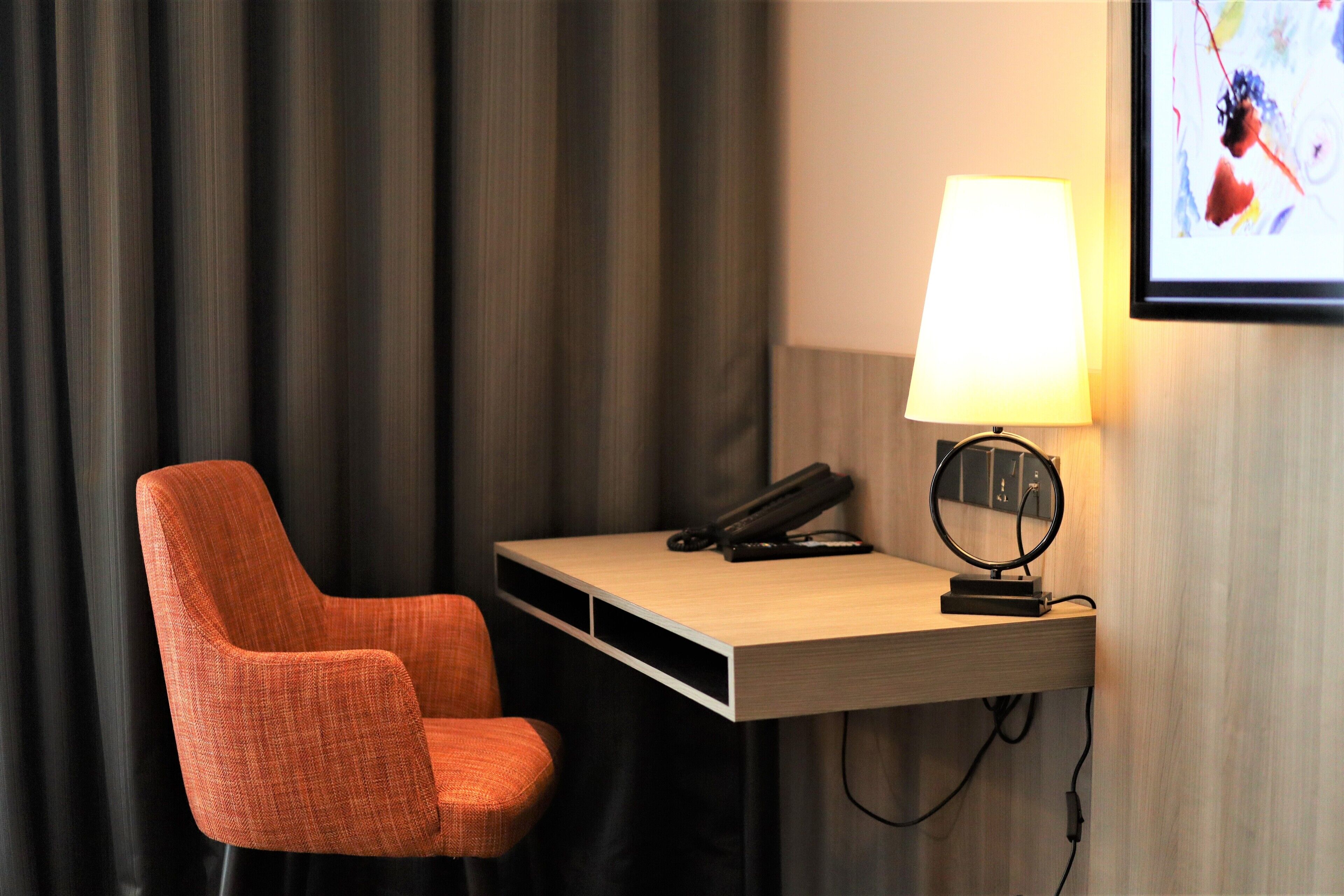 Una habitación de hotel con un escritorio de madera, una silla naranja, una lámpara, un teléfono y un espejo.