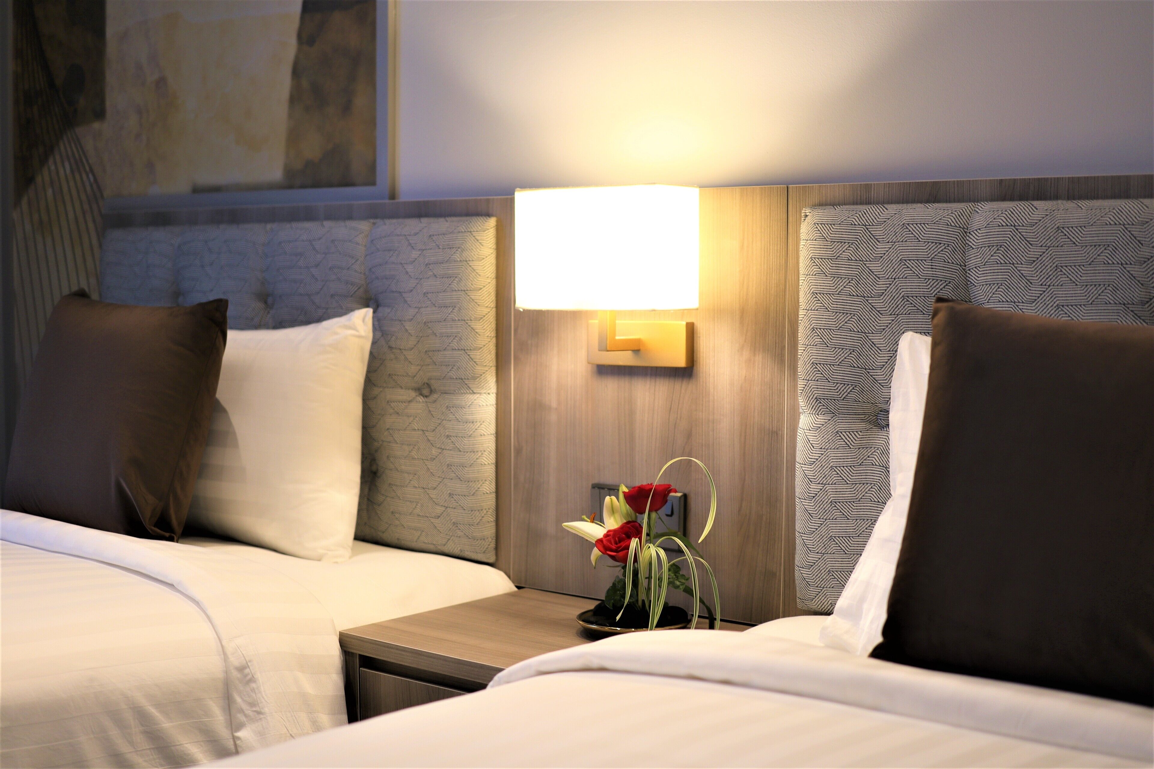 A hotel room with two beds, a nightstand with a lamp, and a vase with flowers.