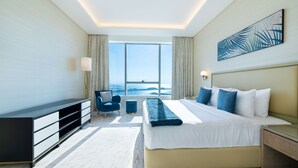 A modern hotel room with a large bed, a desk, a chair, and a view of the sea.