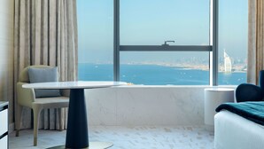 A modern hotel room with a large window offering a view of the sea and a city skyline.
