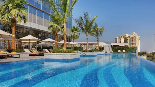 A swimming pool with lounge chairs and umbrellas, surrounded by palm trees and modern buildings.