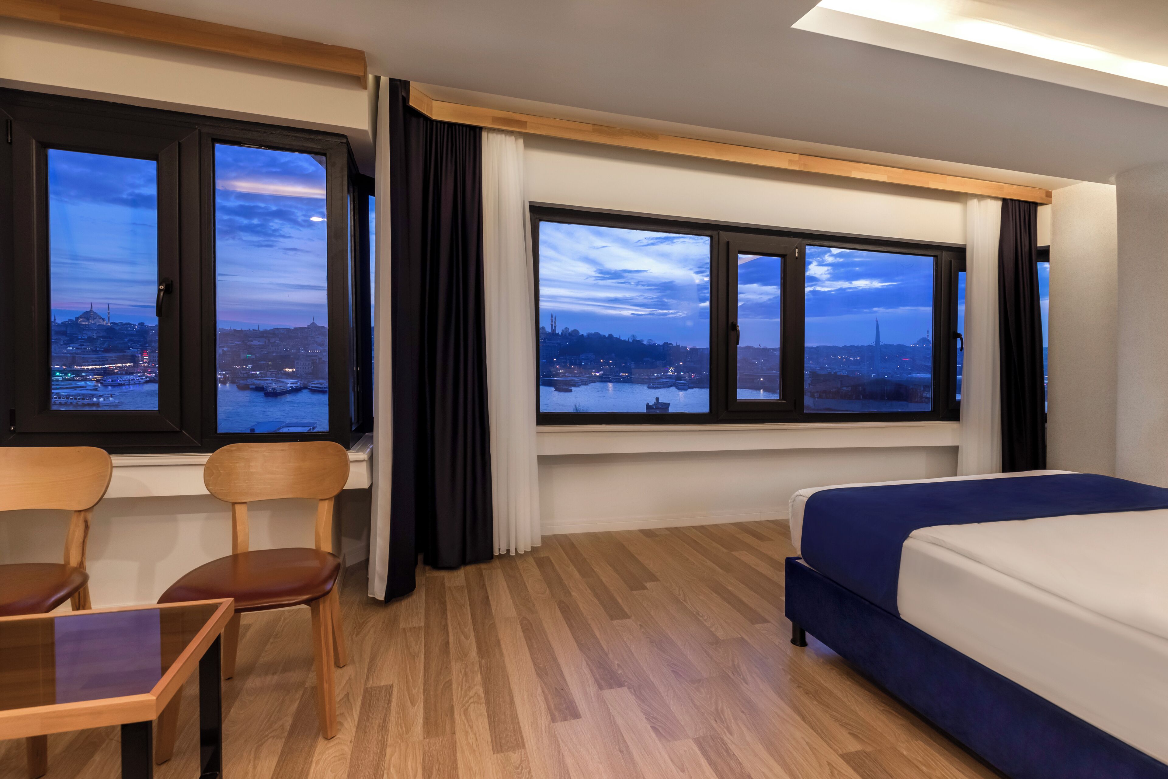 A hotel room with a bed, two chairs, a table, and large windows with a city view.