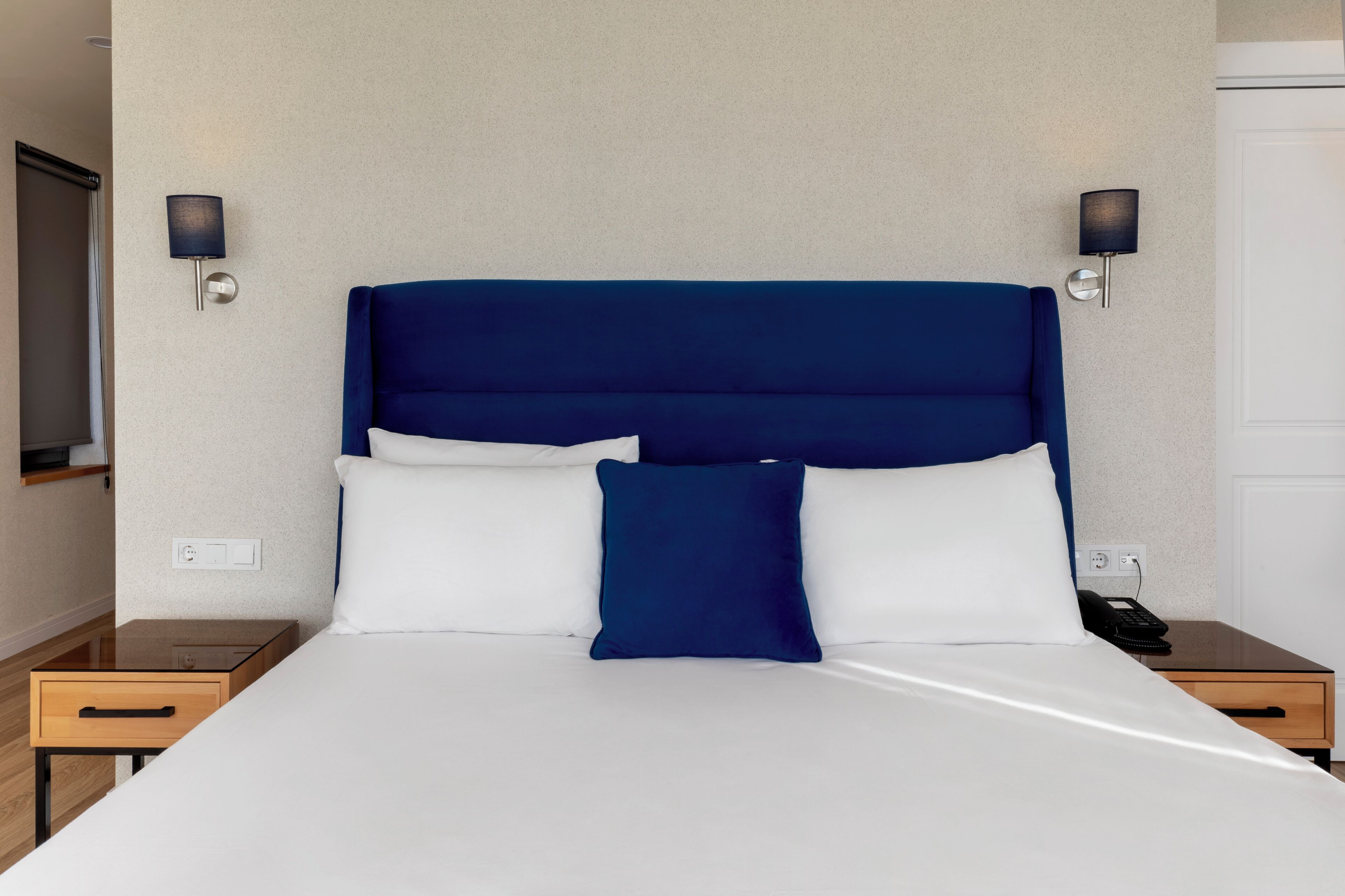 Una habitación de hotel con una cama grande, dos mesitas de noche, un teléfono en la pared y dos apliques.