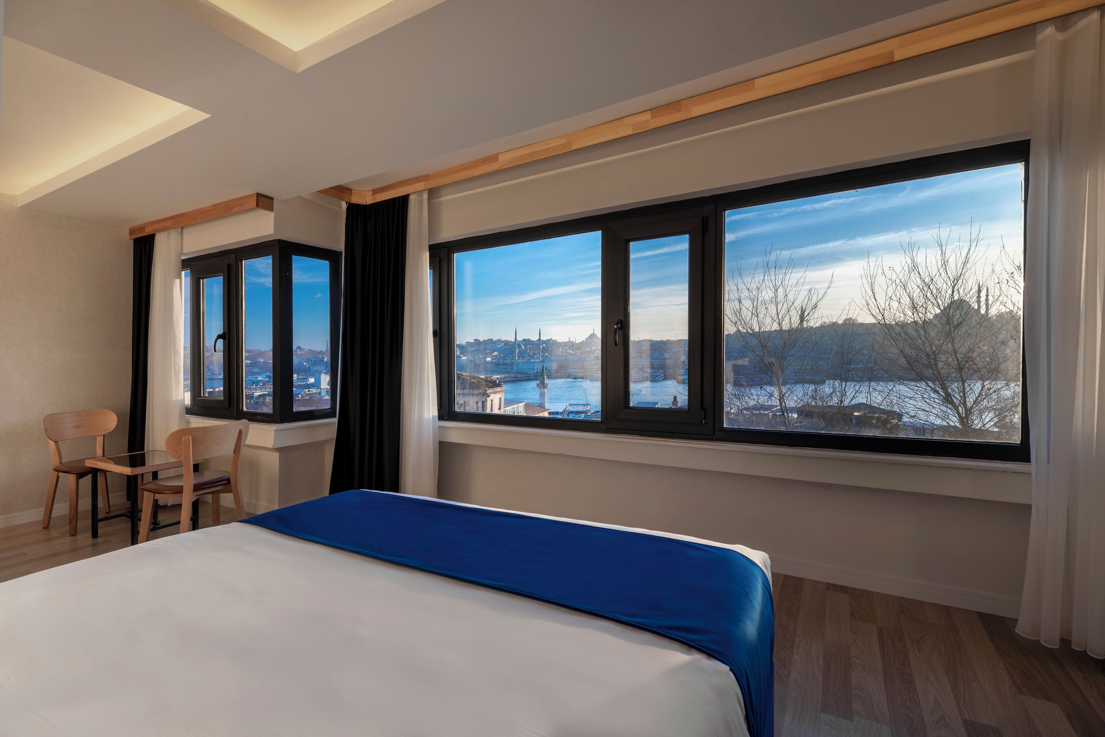 Ein Hotelzimmer mit einem großen Bett, einem Schreibtisch, zwei Stühlen und Blick auf eine Stadtkulisse durch große Fenster.