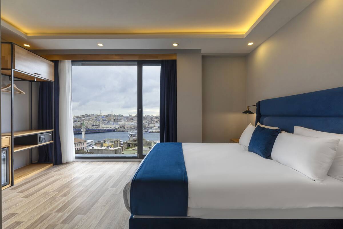 A modern hotel room with a large bed, a wardrobe, and a view of a cityscape through a large window.