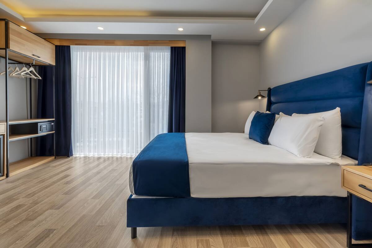 A modern hotel room with a large bed, a wardrobe with hanging clothes, and a large window with curtains.