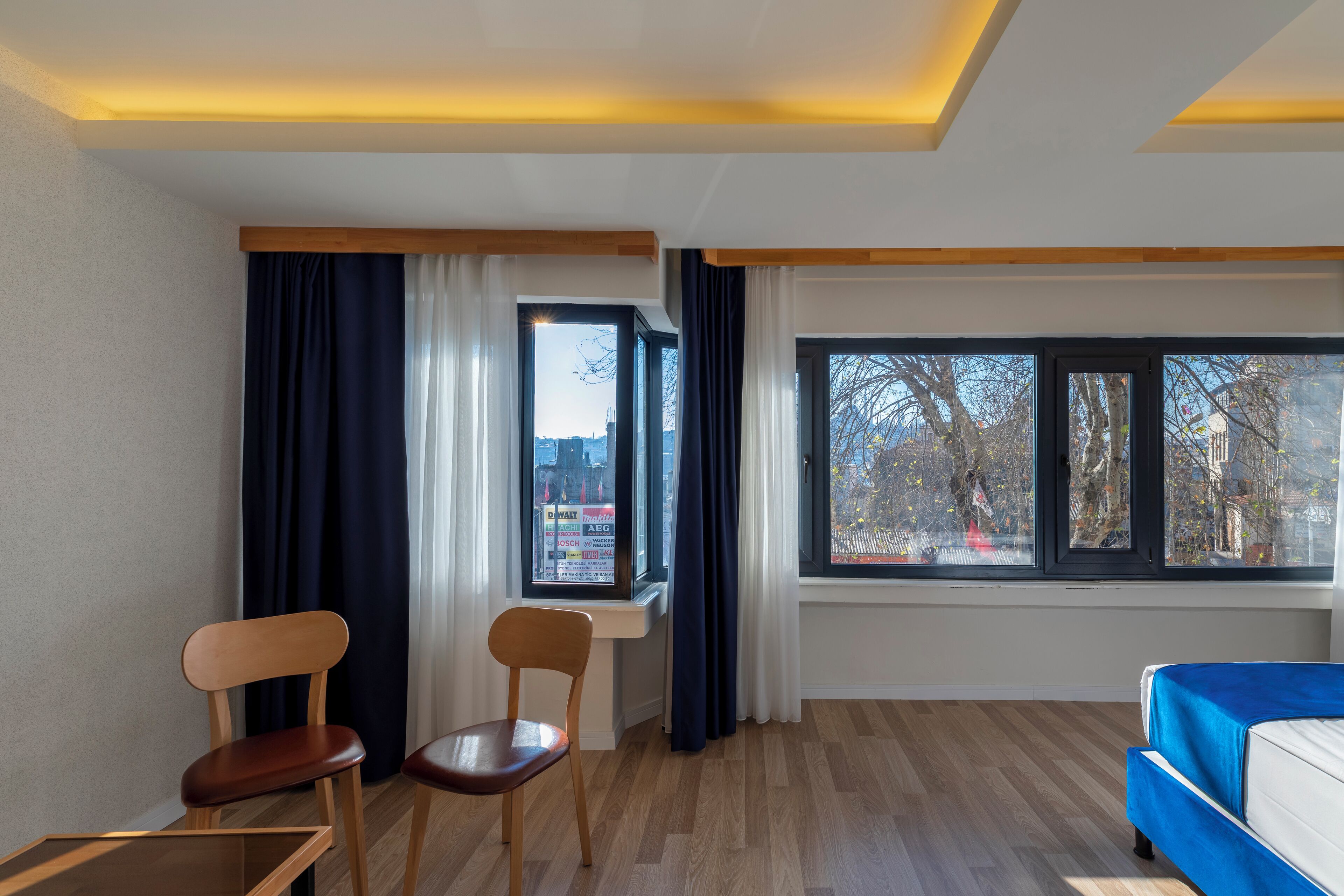 A modern hotel room with a bed, two chairs, a small table, and large windows offering city views.