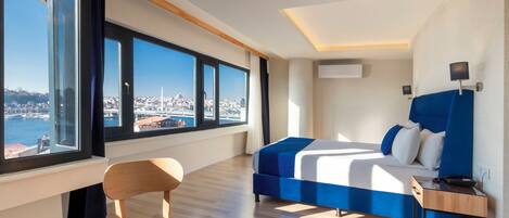 A modern hotel room with a large bed, a desk, and a chair. There is a view of a cityscape through large windows.