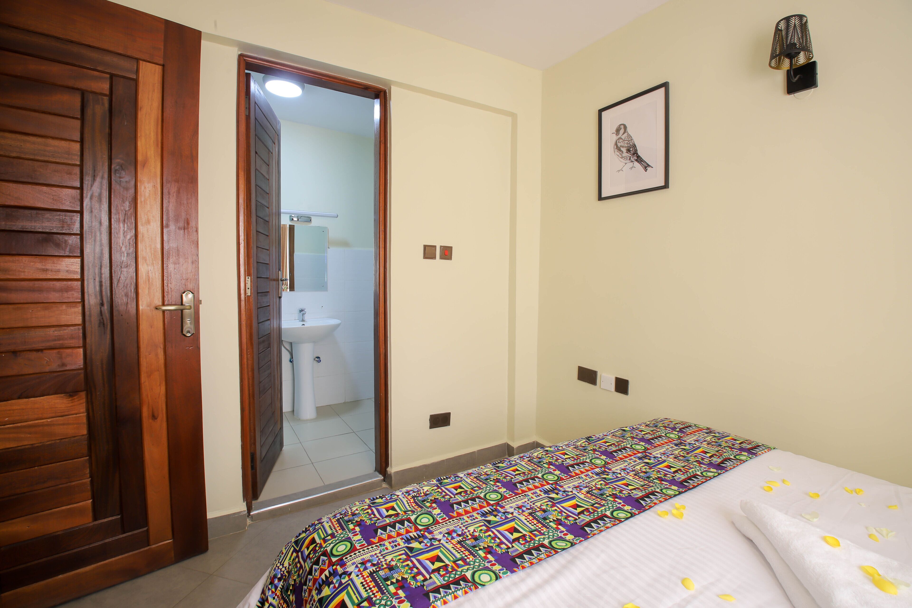 A bedroom with a wooden door, a bed with a colorful blanket, a wall-mounted light fixture, and a framed picture.