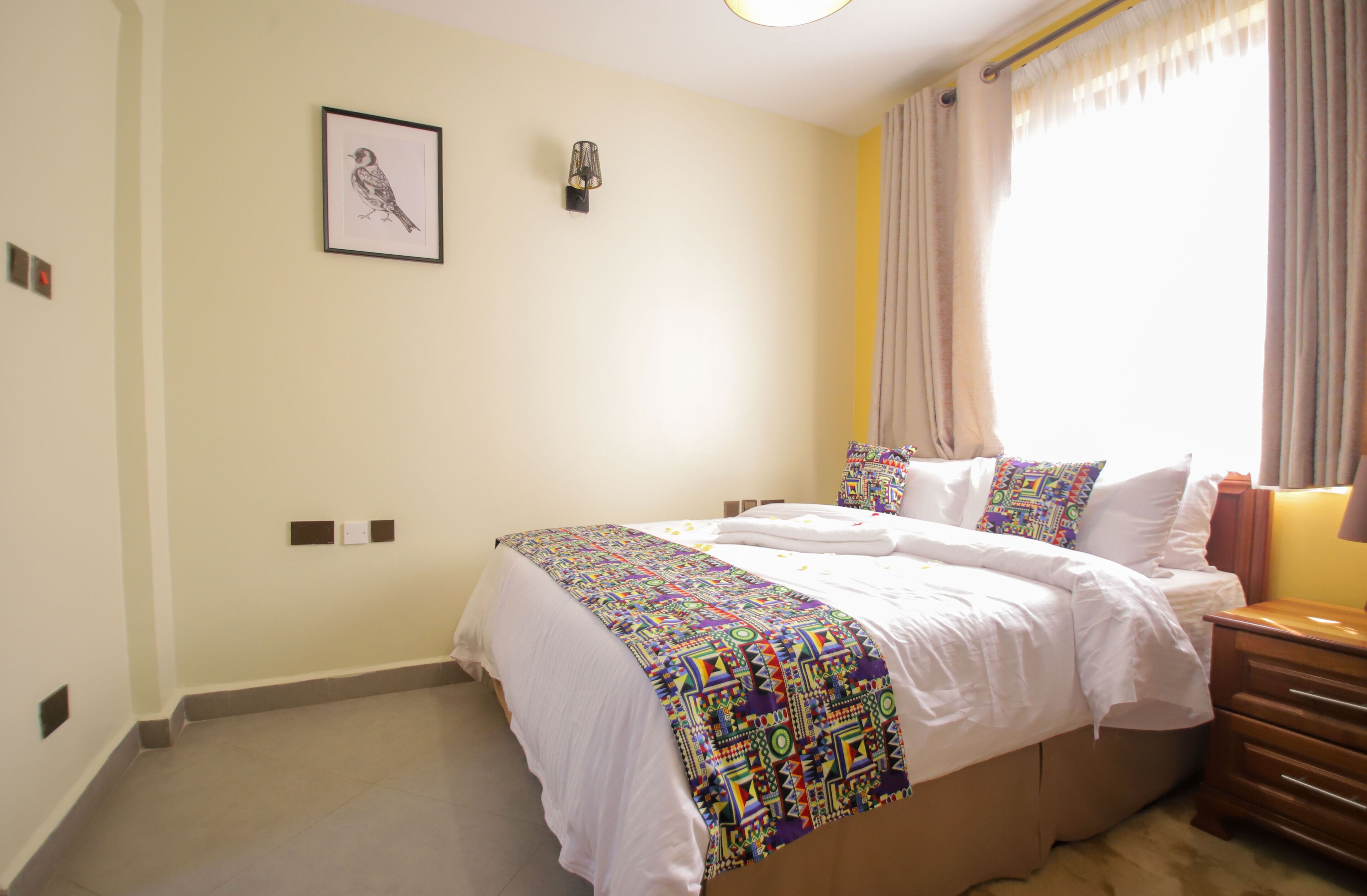 Quarto com cama grande, mesa de cabeceira, luminária na parede e janela com cortinas.