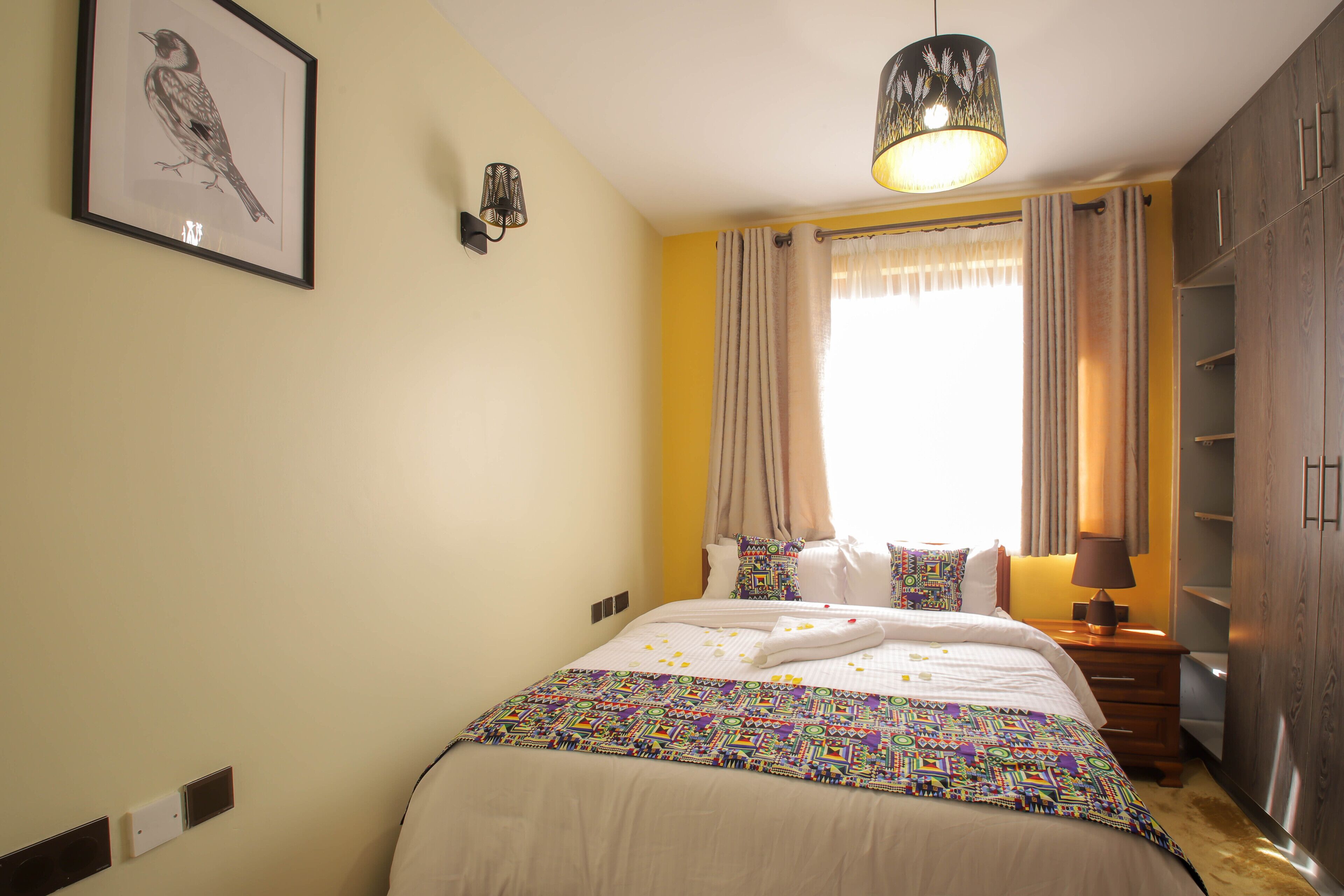 Un letto rifatto con cura, una coperta colorata, un lampadario a parete e un quadro con un uccello.