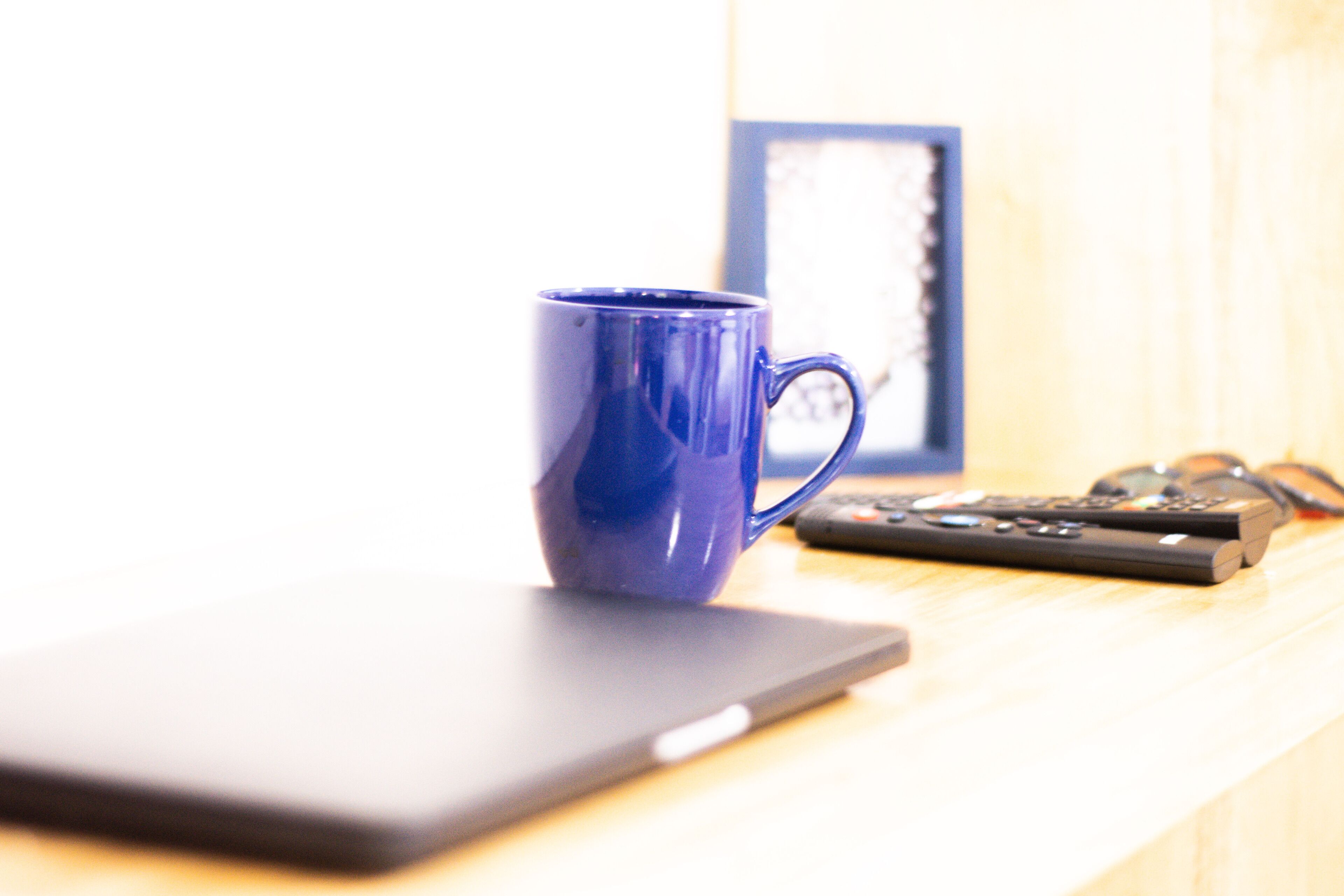 A laptop on a wooden surface with a blue mug and a remote control beside it.