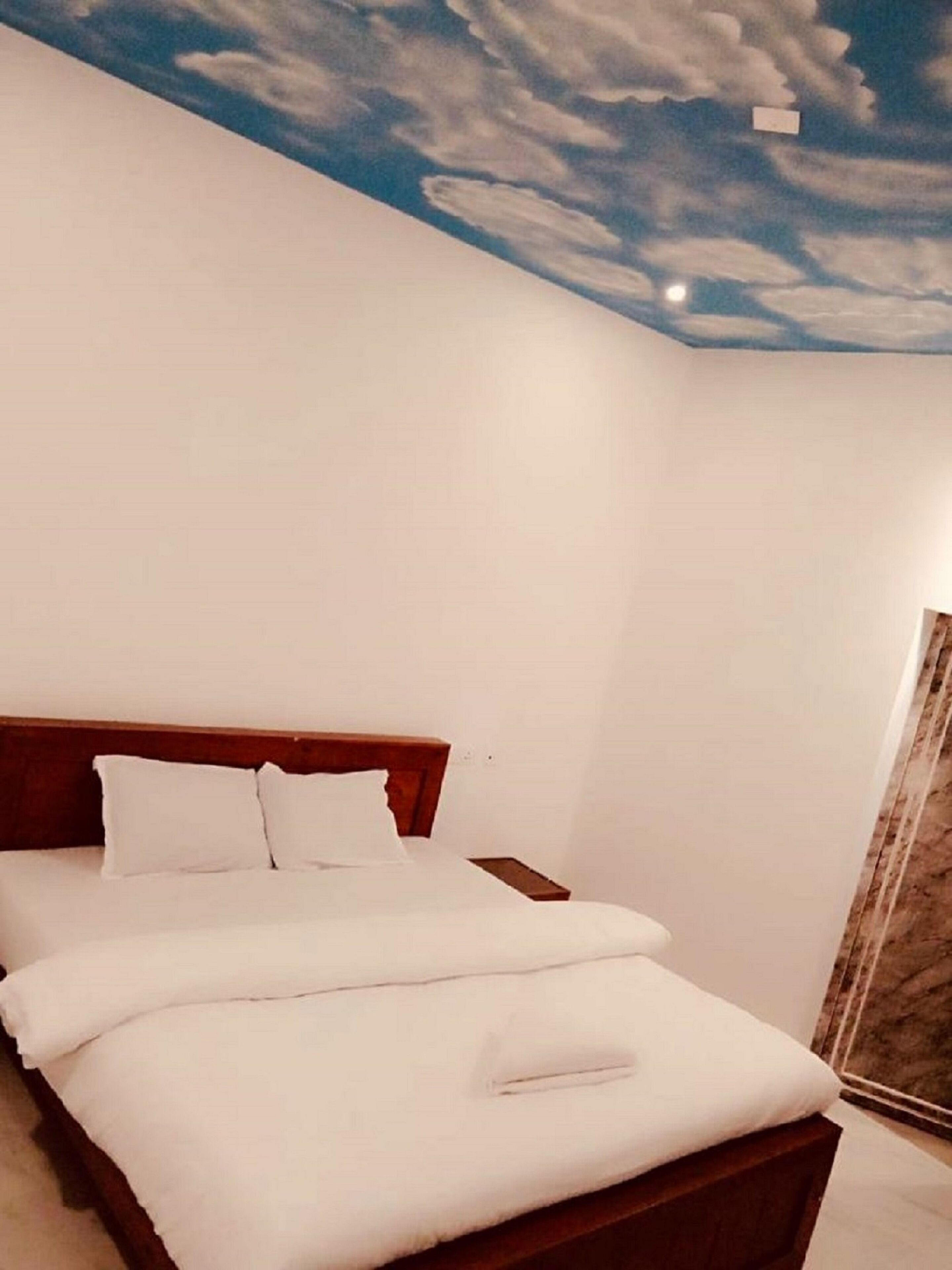 A hotel room with a bed, a wooden headboard, and a ceiling mural of clouds.