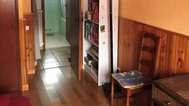 Ett rum med träpanel på väggarna, ett rum som innehåller en säng, ett skrivbord och en stol. Där finns också en bokhylla med diverse föremål och en dörr som leder till ett annat rum.