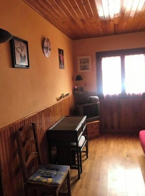 A cozy room with wooden walls and a wooden ceiling, featuring a dining table with chairs, a television, and a window with curtains.